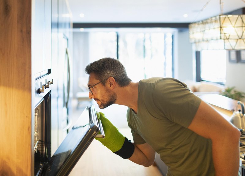 Moški z rokavicami odpira vrata pečice in opazuje jed, ki se peče