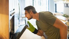 Moški z rokavicami odpira vrata pečice in opazuje jed, ki se peče