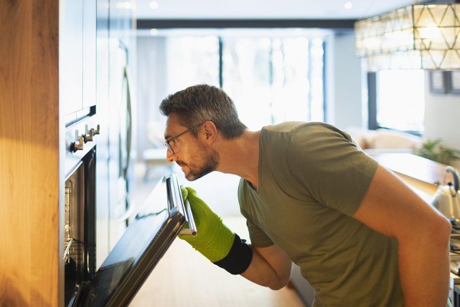 Moški z rokavicami odpira vrata pečice in opazuje jed, ki se peče